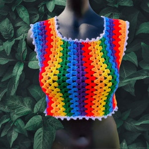 Puede incluir: Un top de crochet arcoíris con un ribete de crochet blanco. El top es sin mangas y tiene un escote redondo.