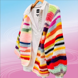 May include: A colorful striped crochet cardigan with long sleeves and a V-neck. The cardigan is made with a variety of bright colors, including pink, red, yellow, green, blue, and purple.