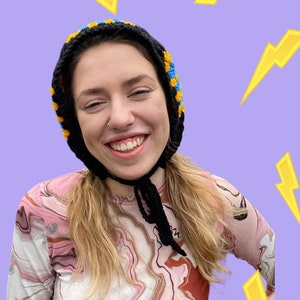 May include: A person wearing a black and blue crocheted hat with a black tie. The person is wearing a pink and brown marbled long-sleeved shirt. The background is a light purple with yellow lightning bolts.