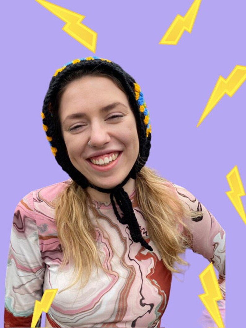 May include: A person wearing a black and blue crocheted hat with a black tie. The person is wearing a pink and brown marbled long-sleeved shirt. The background is a light purple with yellow lightning bolts.