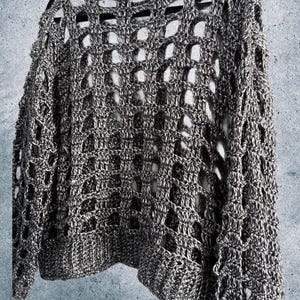 May include: A gray and white open-weave crochet sweater hangs on a black hanger. The sweater has a loose, airy design with large, open spaces throughout the body and sleeves. The texture appears soft and the color is a mix of gray and white.