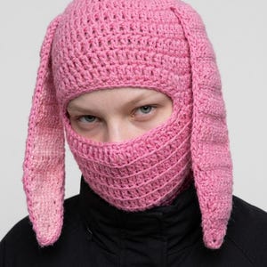 May include: A bright pink crochet balaclava featuring long, dangling rabbit-style ears on both sides. The headgear is constructed with a textured yarn stitch, covering the head and neck while leaving an opening for the eyes and nose area.