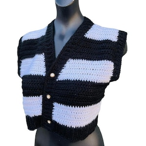 May include: A black and white crochet vest with a V-neck and three gold-colored buttons. The vest features horizontal stripes of black and white, creating a bold, eye-catching pattern. The sleeveless design is displayed on a black mannequin.