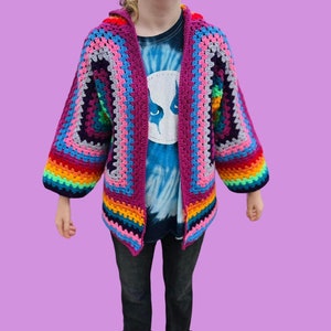 Peut inclure: Un cardigan en crochet arc-en-ciel avec un sch&eacute;ma de couleurs rose, gris et bleu. Le cardigan a une capuche et des manches longues.