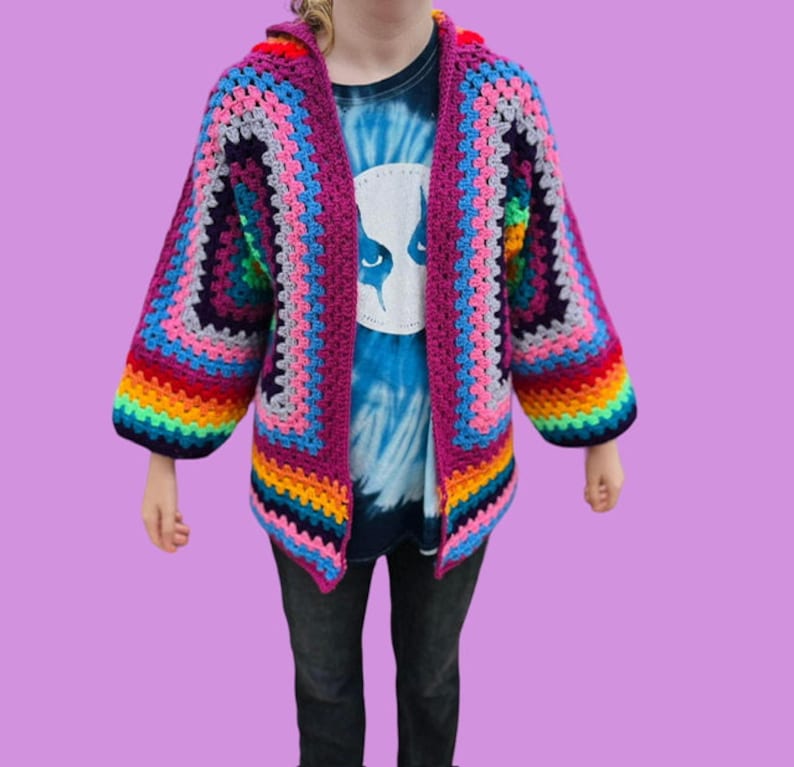 Peut inclure: Un cardigan en crochet arc-en-ciel avec un sch&eacute;ma de couleurs rose, gris et bleu. Le cardigan a une capuche et des manches longues.