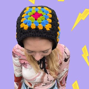 May include: A black crochet hat with a colorful granny square design in yellow, blue, green, and pink. The hat has a long tie that can be worn around the neck.