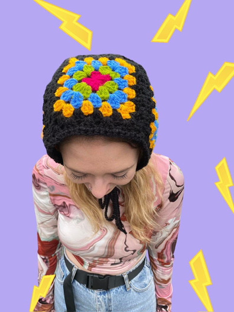 May include: A black crochet hat with a colorful granny square design in yellow, blue, green, and pink. The hat has a long tie that can be worn around the neck.