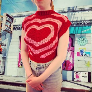 Puede incluir: Un top corto de crochet rojo y rosa con un gran diseño de corazón. El top tiene un cuello alto y mangas cortas.