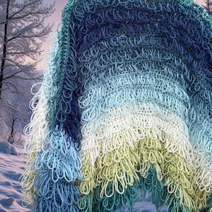 May include: A fringed shawl in a gradient of blues, greens, and white. The shawl is handmade with looped yarn, creating a unique, textured appearance. The colour transitions from dark blue to light blue and white, resembling a winter landscape.