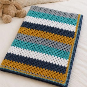 May include: A rectangular crochet blanket featuring horizontal stripes in shades of teal, white, dark blue, mustard yellow, and gray. The blanket has a textured granny square stitch pattern and a dark blue border. A light brown teddy bear is nearby.