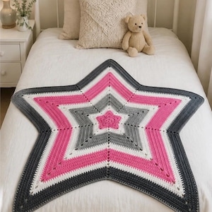 May include: A star-shaped crochet blanket featuring concentric bands of color. The design starts with a pink center, followed by alternating layers of gray, white, and pink, ending with a dark gray border. The texture is consistent throughout.