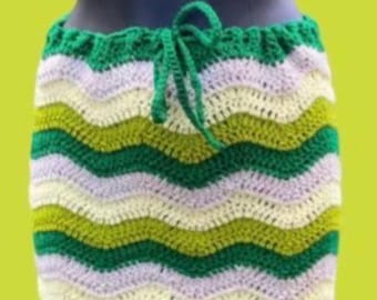 Peut inclure: Jupe au crochet faite à la main avec une taille à cordon verte. La jupe présente un motif en chevrons dans les tons de vert, blanc et violet clair. La jupe est présentée sur un mannequin sur fond jaune-vert.