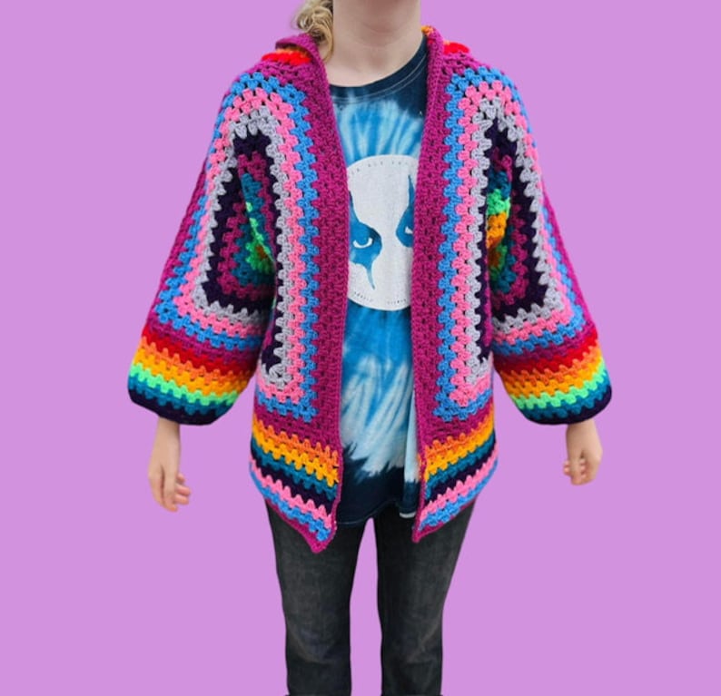 Peut inclure: Un cardigan en crochet color&eacute; avec un motif de rayures arc-en-ciel sur les manches et un corps rose. Le cardigan est ouvert &agrave; l'avant et a une coupe ample.