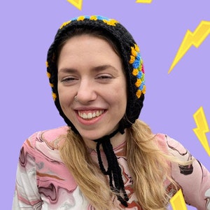 May include: A person wearing a black and colorful crocheted hat with a black cord. The hat has a geometric pattern and is worn over the person's head. The person is wearing a pink and brown patterned shirt.