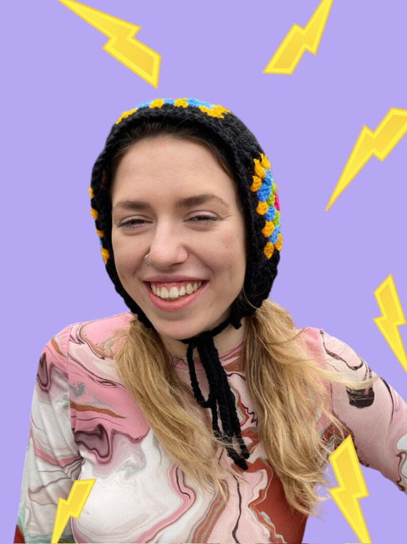 May include: A person wearing a black and colorful crocheted hat with a black cord. The hat has a geometric pattern and is worn over the person's head. The person is wearing a pink and brown patterned shirt.