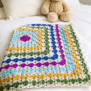 Peut inclure: Une couverture carrée au crochet présentant un motif de granny square répétitif en rangées concentriques de fils beige, bleu clair, jaune, vert, violet et bleu foncé. Un ours en peluche marron clair est posé derrière le textile texturé plié.
