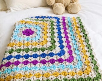 May include: A square crochet blanket featuring a repeating granny square pattern in concentric rows of beige, light blue, yellow, green, purple, and dark blue yarn. A light brown plush teddy bear sits behind the folded, textured textile.