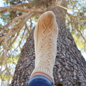 May include: A close-up of a beige knitted sock with a geometric pattern. The sock is worn on a person's foot and the leg is visible in blue jeans.