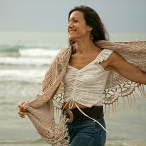 Könnte beinhalten: Eine Frau trägt ein weißes Oberteil mit einem braunen Häkel-Schal, blaue Jeans und einen braunen Gürtel mit einer großen silbernen Schnalle. Sie steht an einem Strand mit dem Meer im Hintergrund.