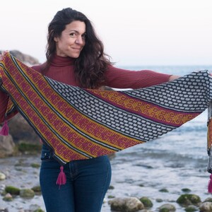Può includere: Una donna con un maglione rosso a collo alto e jeans blu tiene in mano uno scialle di maglia colorato con frange. Lo scialle presenta un motivo geometrico in tonalità di nero, bianco, giallo, rosso e rosa.