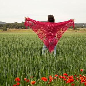 May include: A vibrant red crochet shawl with a floral pattern is displayed against a backdrop of a green wheat field and a cloudy sky. The shawl is triangular and has decorative edging. Red poppies bloom in the foreground.