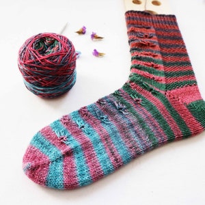 Puede incluir: Un calcetín a rayas de colores con un esquema de color rosa, verde y azul. El calcetín está tejido con un punto texturizado y tiene un agujero en el talón. También se ve en la imagen una bola de hilo con un esquema de color similar.