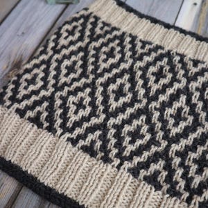 May include: A knitted cowl with a geometric pattern in black and beige. The cowl has a ribbed edge.