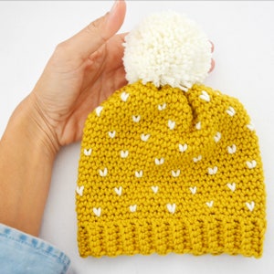 Puede incluir: Un gorro de crochet amarillo con detalles en forma de corazón blanco y un pompón blanco en la parte superior.