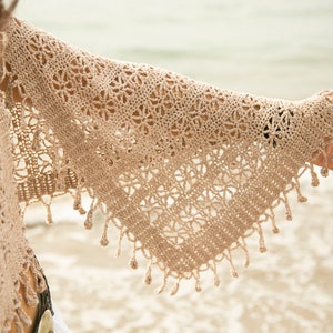 May include: A beige crochet shawl with a delicate lace pattern and fringe trim. The shawl is draped over a person's arm, with the hand extended outward.