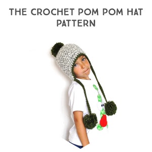 May include: A child wearing a white shirt with a green and red graphic print is wearing a gray and white crocheted hat with a green pom pom on top. The hat has long green crocheted ties with green pom poms on the ends.