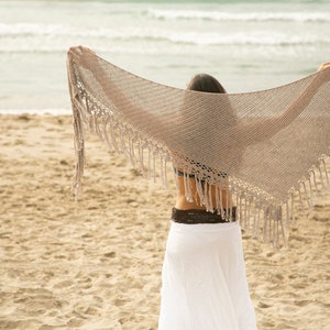 May include: A woman on a sandy beach wearing a white skirt and a crocheted shawl with fringe. The shawl is draped over her shoulders and she is holding the edges of the shawl in her hands.