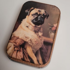 May include: A vintage-style tin box with a brown and white pug dog wearing a lace collar and dress. The dog is sitting on a brown background with a vase of flowers.