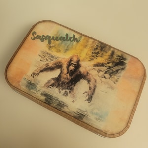 May include: A vintage-style tin box with a watercolor illustration of a Sasquatch in a forest setting. The text "Sasquatch" is written in green lettering on the box.