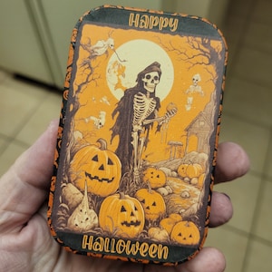 May include: A small, rectangular tin with a Halloween theme. The tin features a skeleton in a black robe holding a pumpkin, surrounded by pumpkins and a spooky house. The text "Happy Halloween" is printed on the top of the tin.
