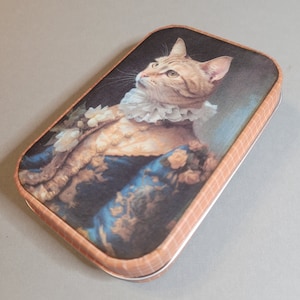 May include: A vintage-style tin box with a portrait of a cat wearing a ruffled collar and a blue and gold patterned dress. The box has a checkered pattern on the lid.