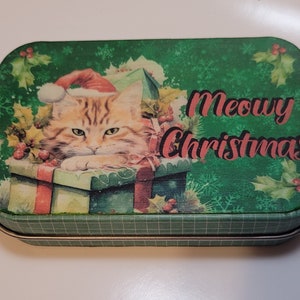 May include: A green tin box with a Christmas cat design. The cat is wearing a Santa hat and is sitting in a box with presents. The text on the box says "Meowy Christmas".