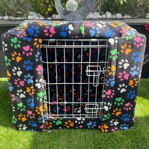 May include: A black pet crate cover featuring a repeating pattern of colourful paw prints. The cover is designed to fit over a wire crate, providing a cosy space for pets. The paw prints are in various colours.