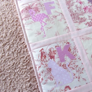 May include: A pink and white patchwork quilt with a toile pattern and appliqued letters and shapes. The quilt features a flamingo, cat, and other shapes. The background is a soft, textured pink fabric.