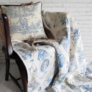 May include: A blue and white toile patterned cushion and quilt set. The cushion has a black trim. The quilt is draped over a wooden chair. The toile design depicts pastoral scenes. The chair is made of wood.