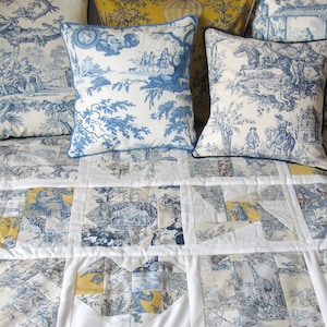 Könnte beinhalten: Eine weiße Steppdecke mit einem blauen und gelben Toile-Muster. Die Steppdecke besteht aus Quadraten und ist mit mehreren blauen und weißen gemusterten Kissen verziert.