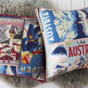 May include: Two square cushions with vintage travel poster designs. One shows a Swiss mountain scene with "SWITZERLAND". The other says "AUSTRIA" and "Visit AUSTRIA To Ski!" in red and blue. Both cushions have a red trim and are on a grey faux fur surface.