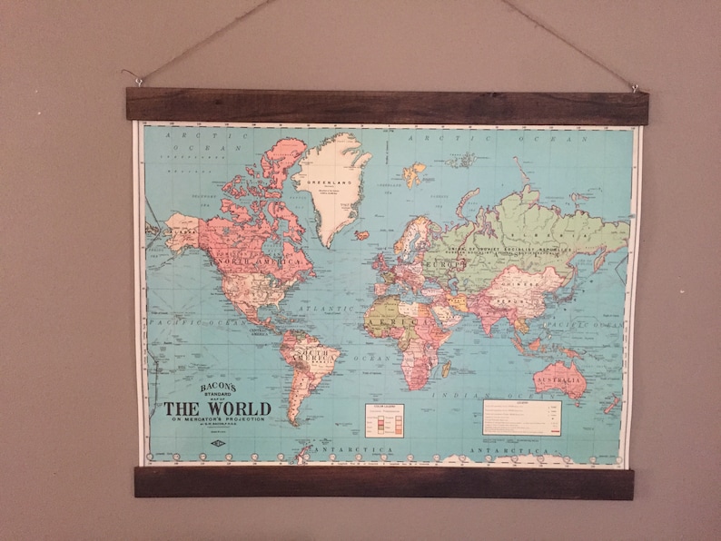 Pu&ograve; includere: Una mappa del mondo vintage con sfondo blu e continenti in vari colori. La mappa &egrave; incorniciata in una cornice di legno scuro ed &egrave; appesa a un muro. La mappa &egrave; intitolata "Bacon's Standard The World On Mercator's Projection".