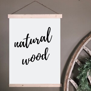 May include: A white poster with the words "natural wood" in black script lettering. The poster is held by two natural wood strips and suspended by a twine string. The background is a neutral gray.
