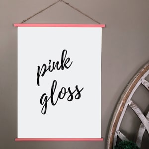 May include: A white poster with the words "pink gloss" in black script, held by pink wooden bars and a twine hanger. The poster is displayed against a neutral gray wall, creating a clean and modern aesthetic.