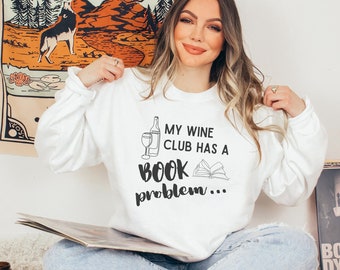 May include: White long-sleeved sweatshirt with the text "MY WINE CLUB HAS A BOOK problem..." in black. The graphic includes a wine bottle, a wine glass, and an open book. The person is holding a vinyl record.