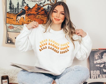 May include: White long-sleeved sweatshirt with the text "READ BANNED BOOKS" in a curved, layered design of orange and yellow. The sweatshirt is worn by a person holding a record album.