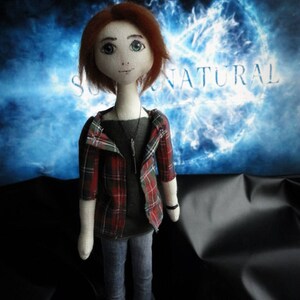 Puede incluir: Mu&ntilde;eca hecha a mano con cabello rojo, que lleva una camisa gris, vaqueros azules y una chaqueta a cuadros roja y verde. La mu&ntilde;eca tiene un collar y zapatos negros. El fondo presenta la palabra "SUPERNATURAL" en blanco sobre un dise&ntilde;o azul y blanco.