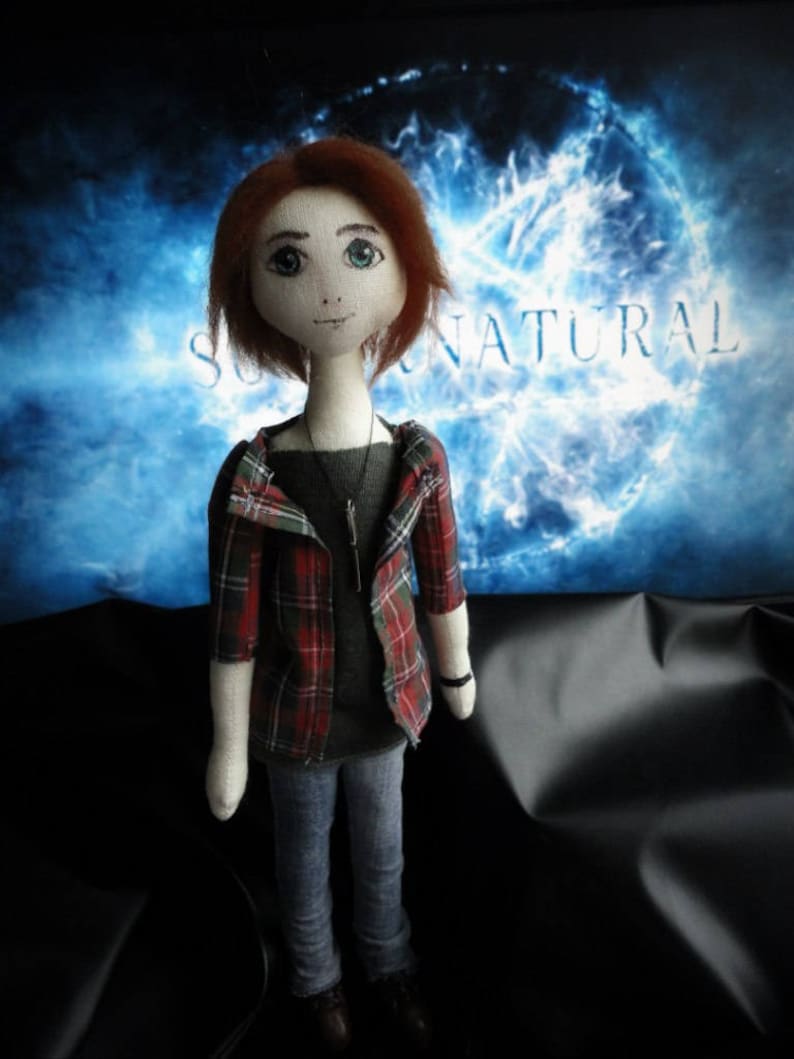 Puede incluir: Mu&ntilde;eca hecha a mano con cabello rojo, que lleva una camisa gris, vaqueros azules y una chaqueta a cuadros roja y verde. La mu&ntilde;eca tiene un collar y zapatos negros. El fondo presenta la palabra "SUPERNATURAL" en blanco sobre un dise&ntilde;o azul y blanco.