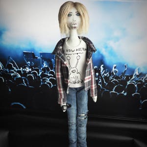 Puede incluir: Mu&ntilde;eca hecha a mano vestida con una camisa a cuadros, vaqueros rotos y una camiseta blanca con un gr&aacute;fico. La mu&ntilde;eca tiene el pelo rubio y sostiene un micr&oacute;fono. El fondo representa una escena de concierto.