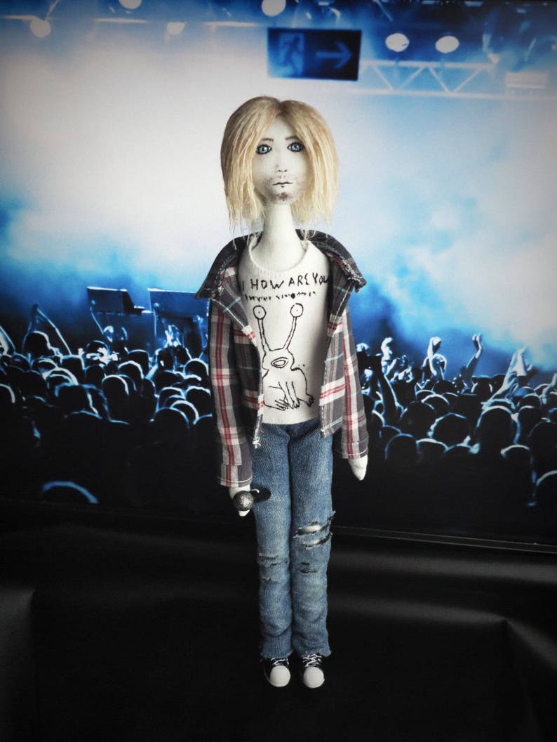 Puede incluir: Mu&ntilde;eca hecha a mano vestida con una camisa a cuadros, vaqueros rotos y una camiseta blanca con un gr&aacute;fico. La mu&ntilde;eca tiene el pelo rubio y sostiene un micr&oacute;fono. El fondo representa una escena de concierto.